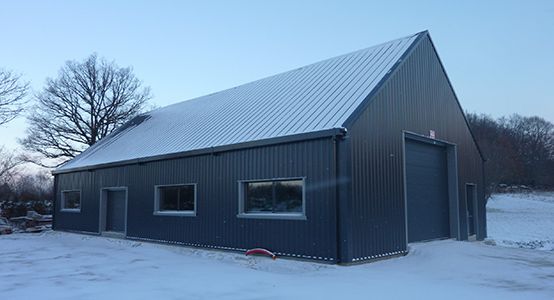 Grange en métal gris, toit et sol enneigés, se détachant sur un arbre dénudé et un ciel couvert.