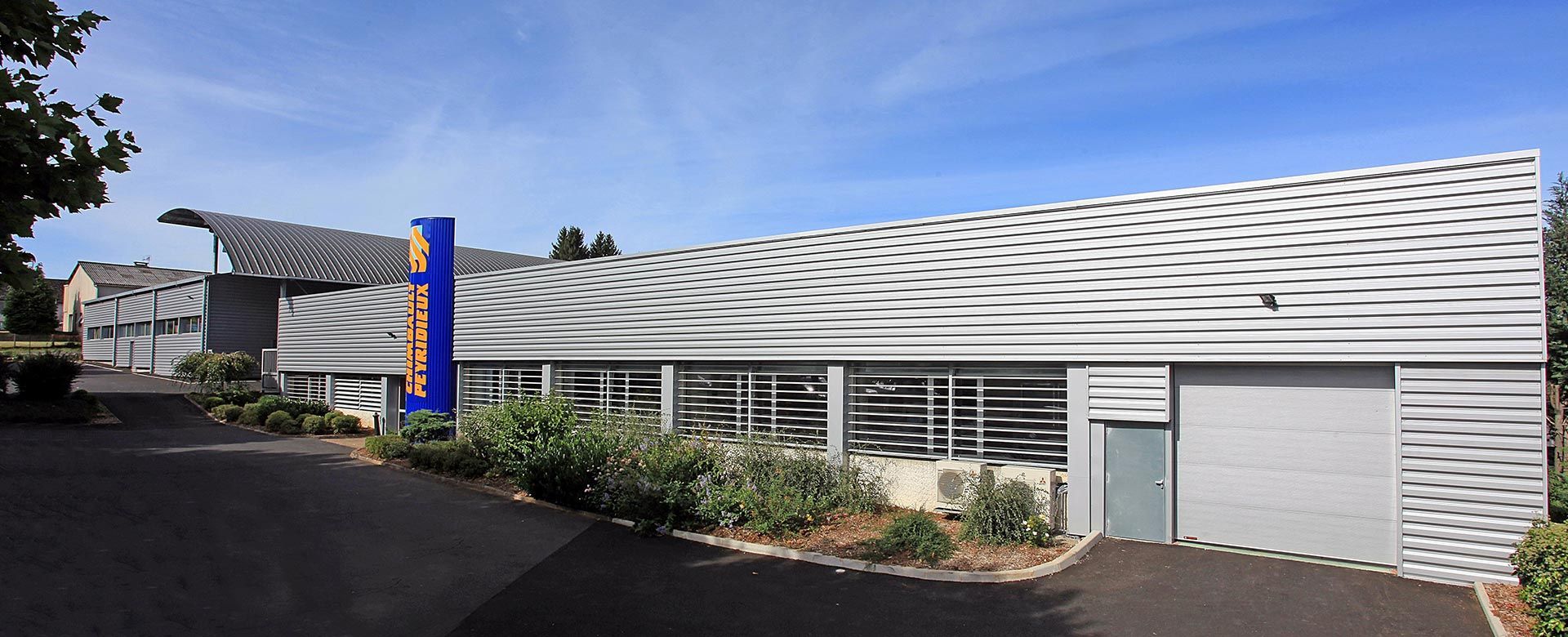 Bâtiment moderne avec une façade à motifs ondulés blancs et gris, enseigne bleue, porte bleue et asphalte noir.