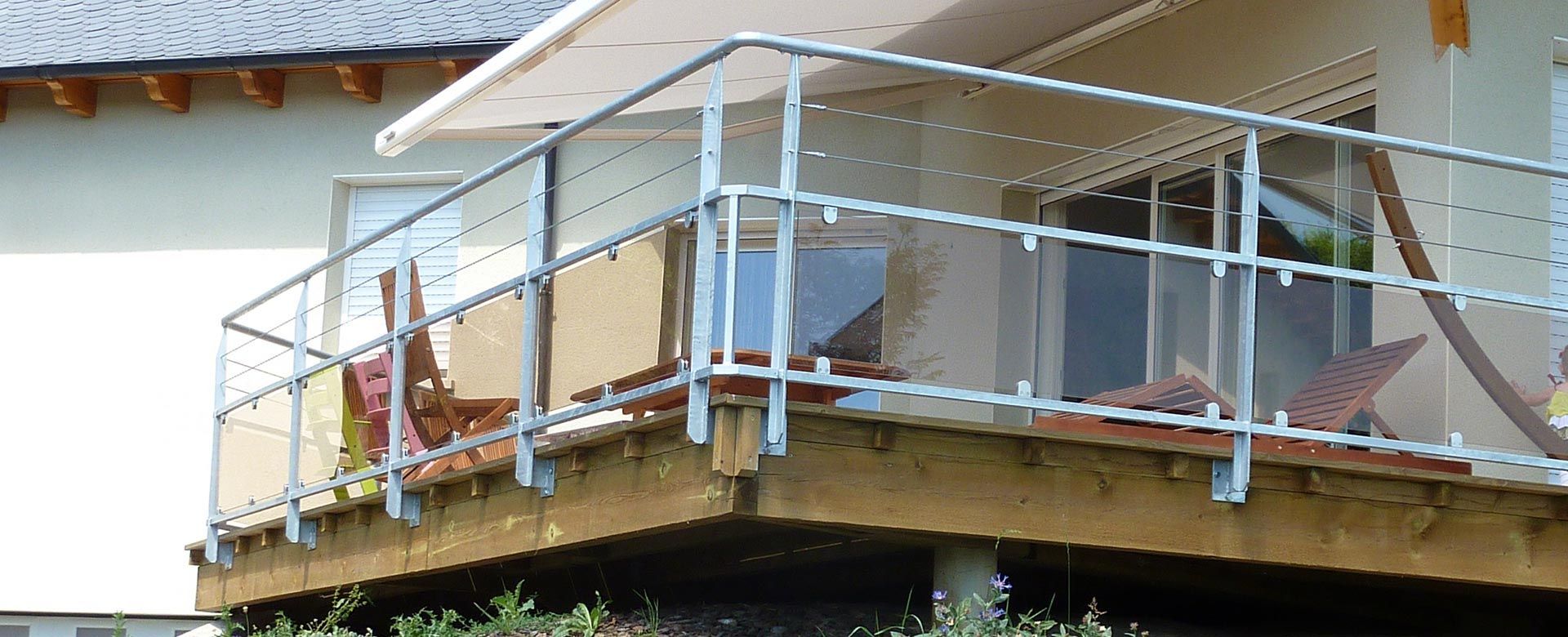 Une terrasse en bois avec une rambarde en verre. Une maison partiellement visible se trouve à l'arrière-plan.