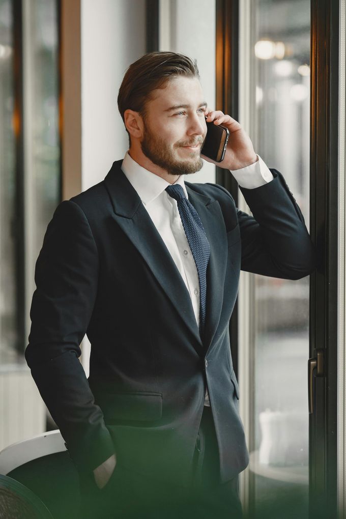 Un hombre con traje oscuro habla por teléfono inteligente junto a una ventana en el vestíbulo de una oficina.