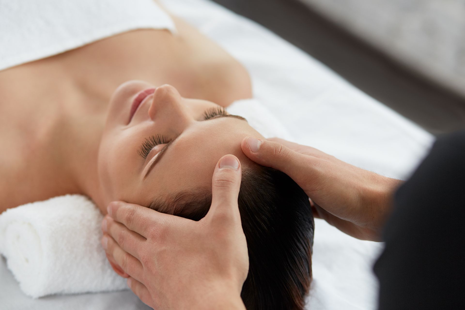 Une femme reçoit un massage du visage, les mains posées sur le front, sur une table de massage.