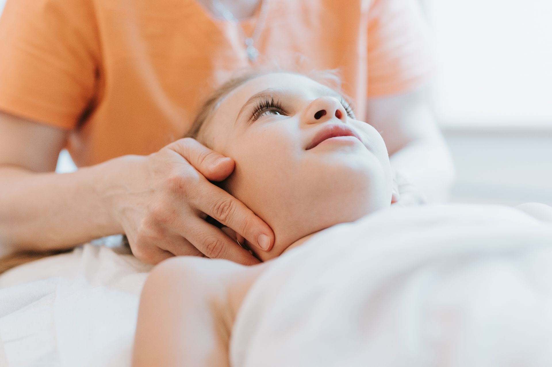 Des mains massent le visage d'un enfant allongé sur une surface blanche. L'enfant regarde vers le ciel.