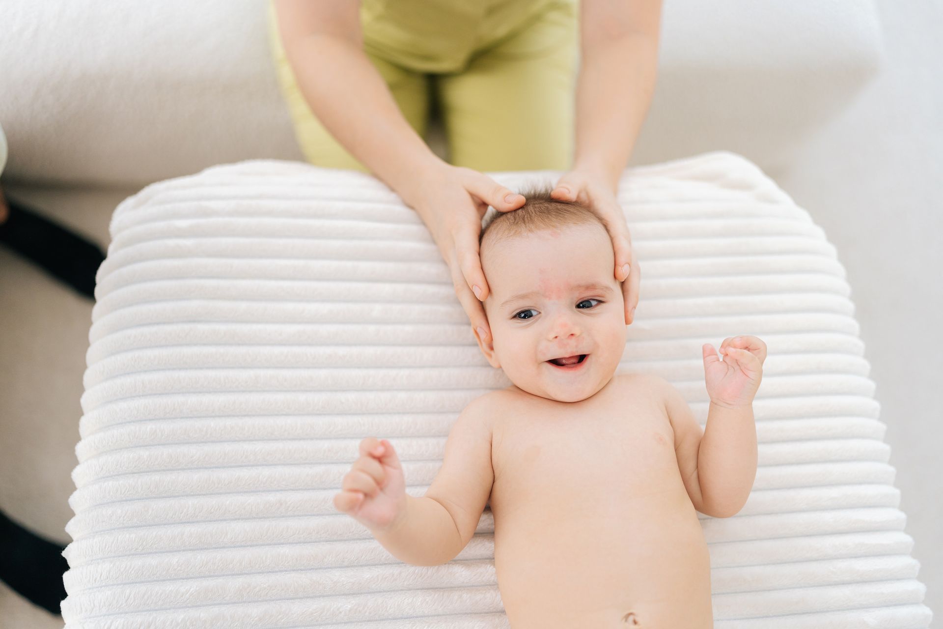 Un bébé, allongé sur une couverture blanche, sourit aux mains d'une personne qui lui caressent doucement la tête.