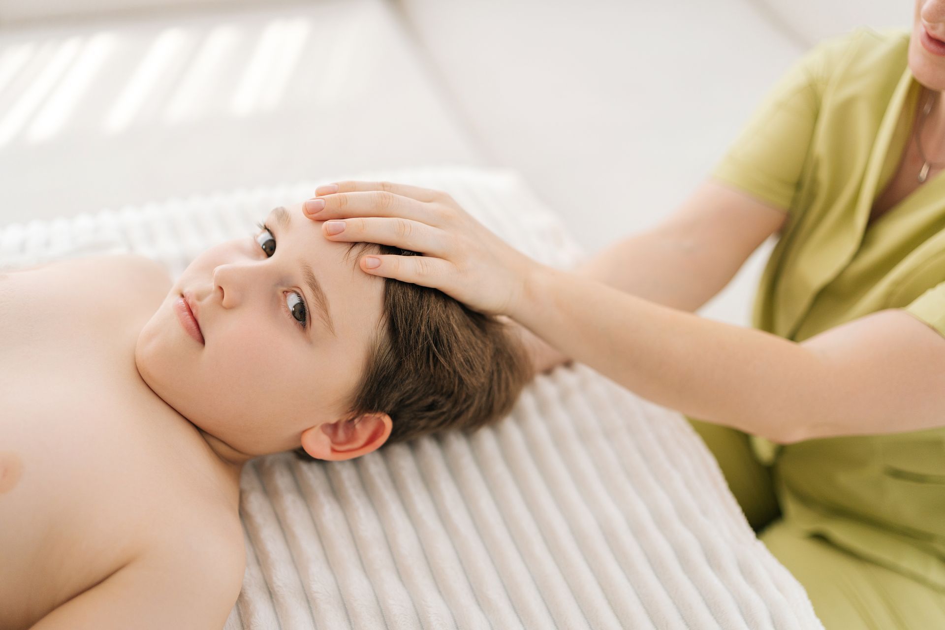 Un enfant reçoit un massage de la tête ; la main d'une personne est posée sur son front. L'enfant est allongé sur une surface blanche, torse nu ; la personne est vêtue de vert.