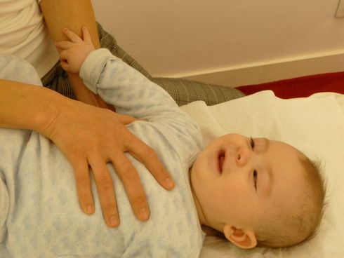 Un bébé allongé sur une surface blanche, tenu et touché par les mains d'une personne.