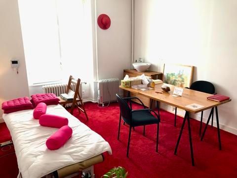 Chambre avec table de massage, bureau, chaises et tapis rouge. Coussins rose vif et chapeau rouge.