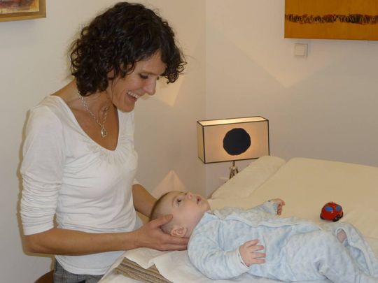 Une femme sourit à un bébé allongé sur un lit blanc. Elle tient doucement la tête de l'enfant. Chambre lumineuse.