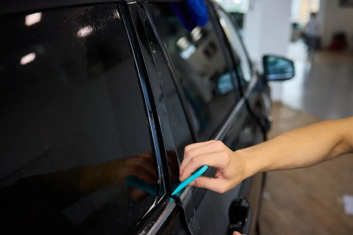 Persona que utiliza una herramienta azul para recortar el tinte de la ventana de una puerta de automóvil negra en un garaje.