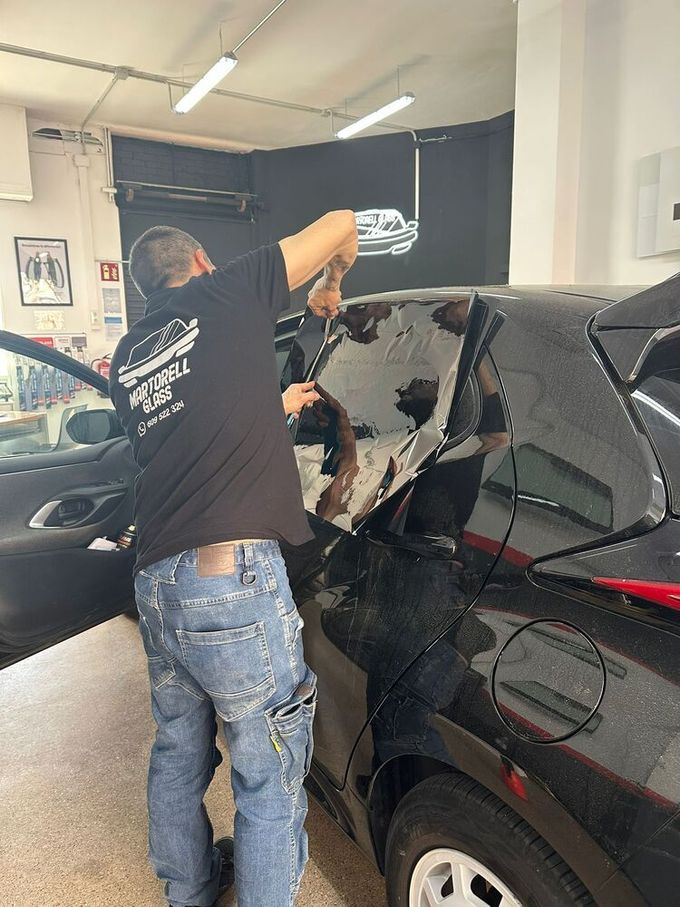Persona que utiliza una herramienta azul para recortar el tinte de la ventana de una puerta de automóvil negra en un garaje.