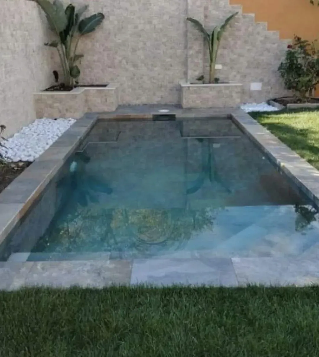 Pequeña piscina rectangular con borde de azulejos grises, escalones y agua cristalina; flanqueada por jardineras y césped.