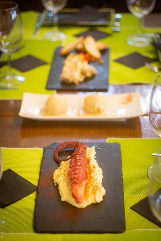 Aperitivos en la mesa: pulpo y puré de patatas, comida frita en pizarra y comida esférica en un plato largo.