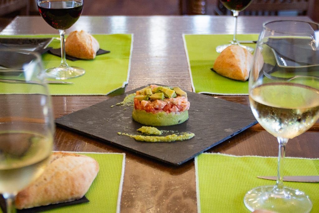 Una comida en una mesa con copas de vino, pan y un plato con ingredientes verdes y rojos.