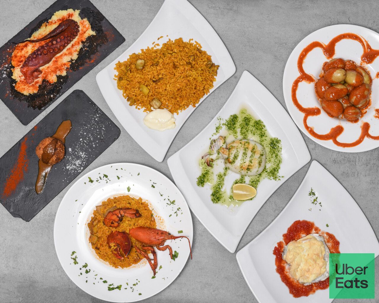 Platos variados de mariscos en platos blancos, que incluyen paella y calamares a la parrilla, dispuestos sobre una superficie gris, con el logotipo de Uber Eats visible.