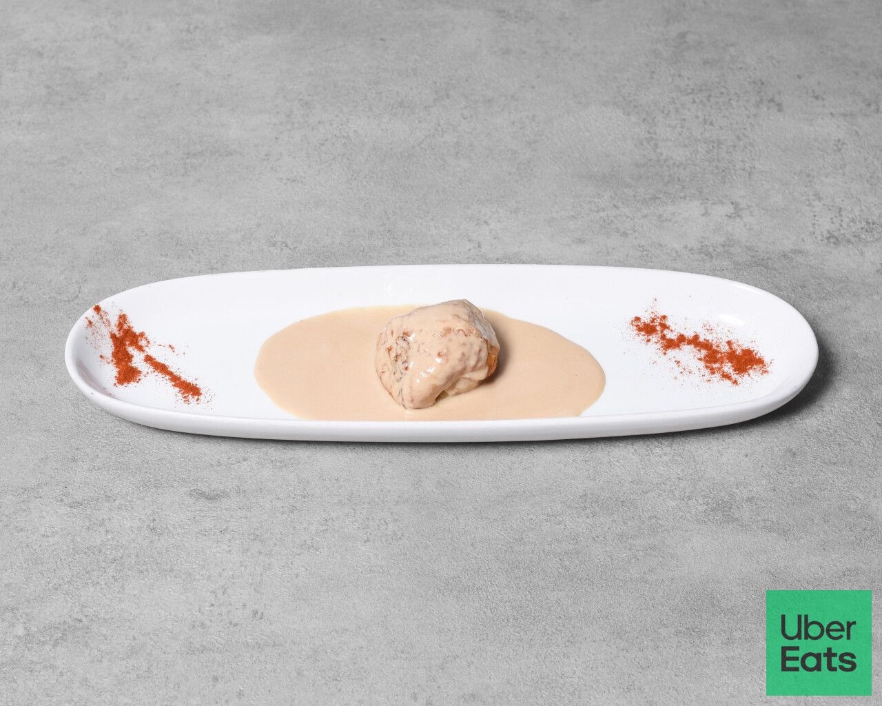 Un alimento pequeño cocinado en un plato ovalado blanco con salsa y guarnición roja, sobre una superficie gris.