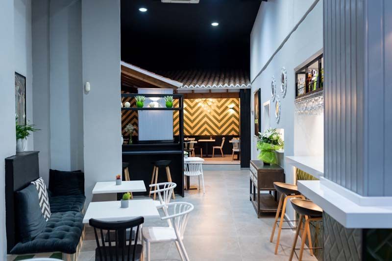 Interior de un café moderno: paredes grises, techo negro, mesas, sillas, barra y paneles decorativos de madera en la parte trasera.