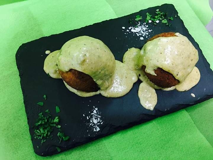 Dos porciones redondas de comida frita sobre una pizarra oscura, cubiertas con salsa cremosa y adornadas con hierbas picadas.