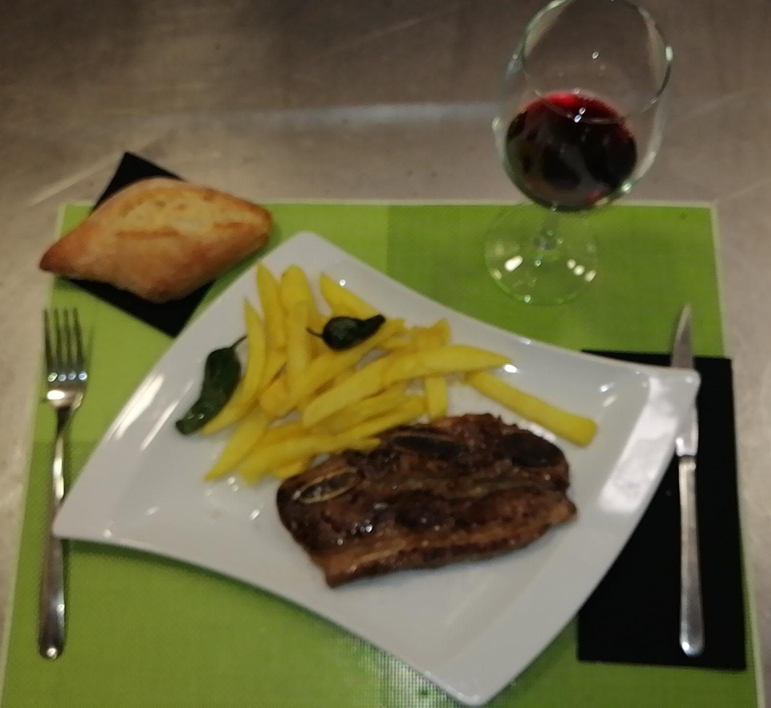 Bistec, patatas fritas, pan y vino en un plato con cubiertos, servido sobre un mantel verde y negro.