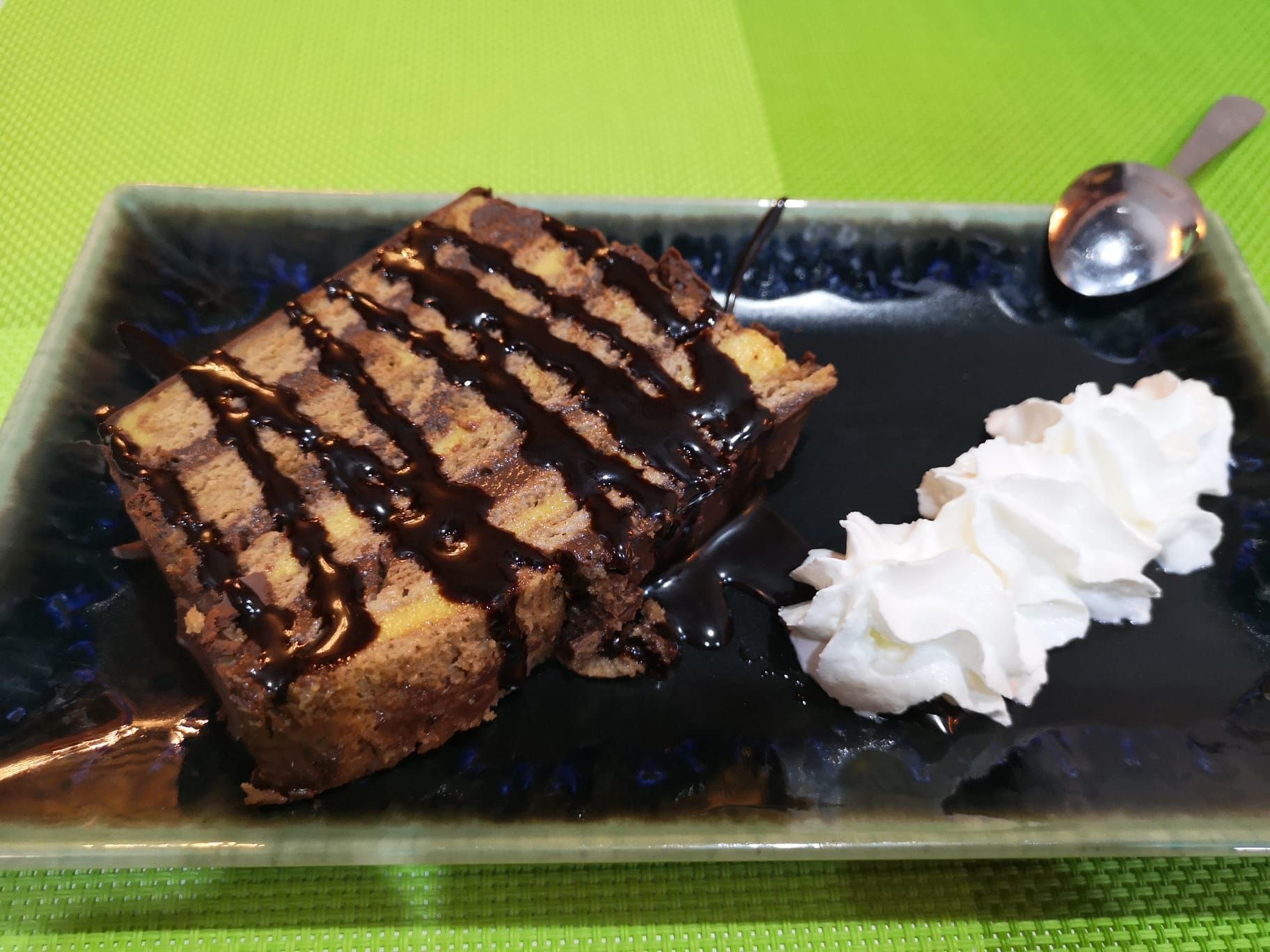 Rebanada de pastel de chocolate rociada con salsa de chocolate y crema batida en un plato oscuro con una cuchara.