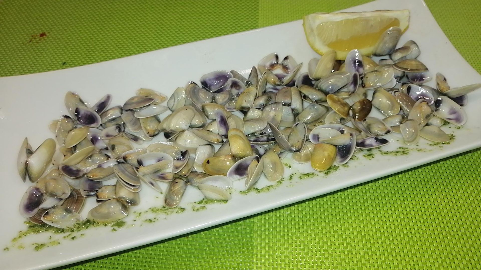 Almejas servidas en un plato rectangular blanco con una rodaja de limón y un mantel verde.
