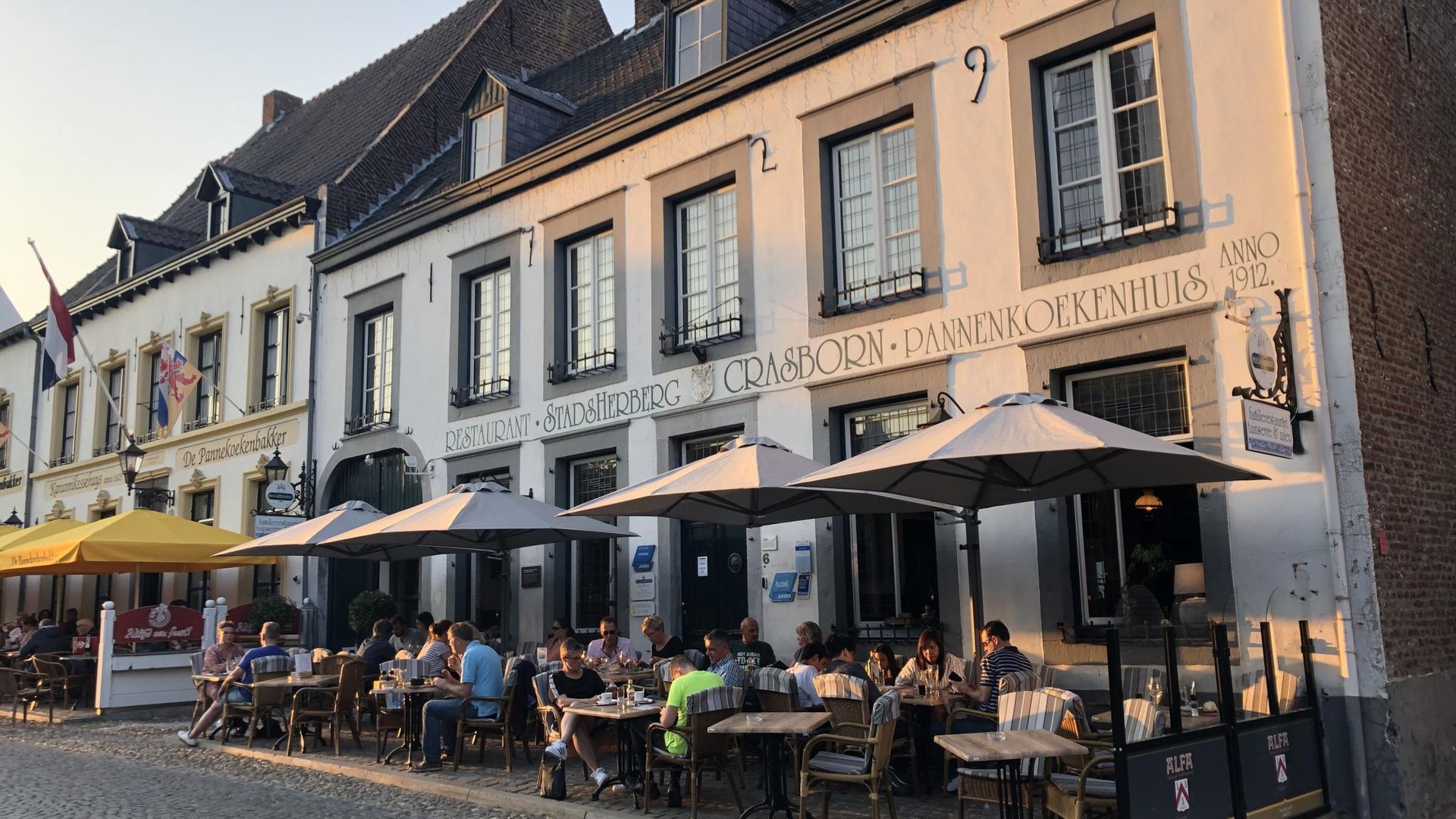 Een groep mensen zit aan tafels buiten een gebouw, met parasols.