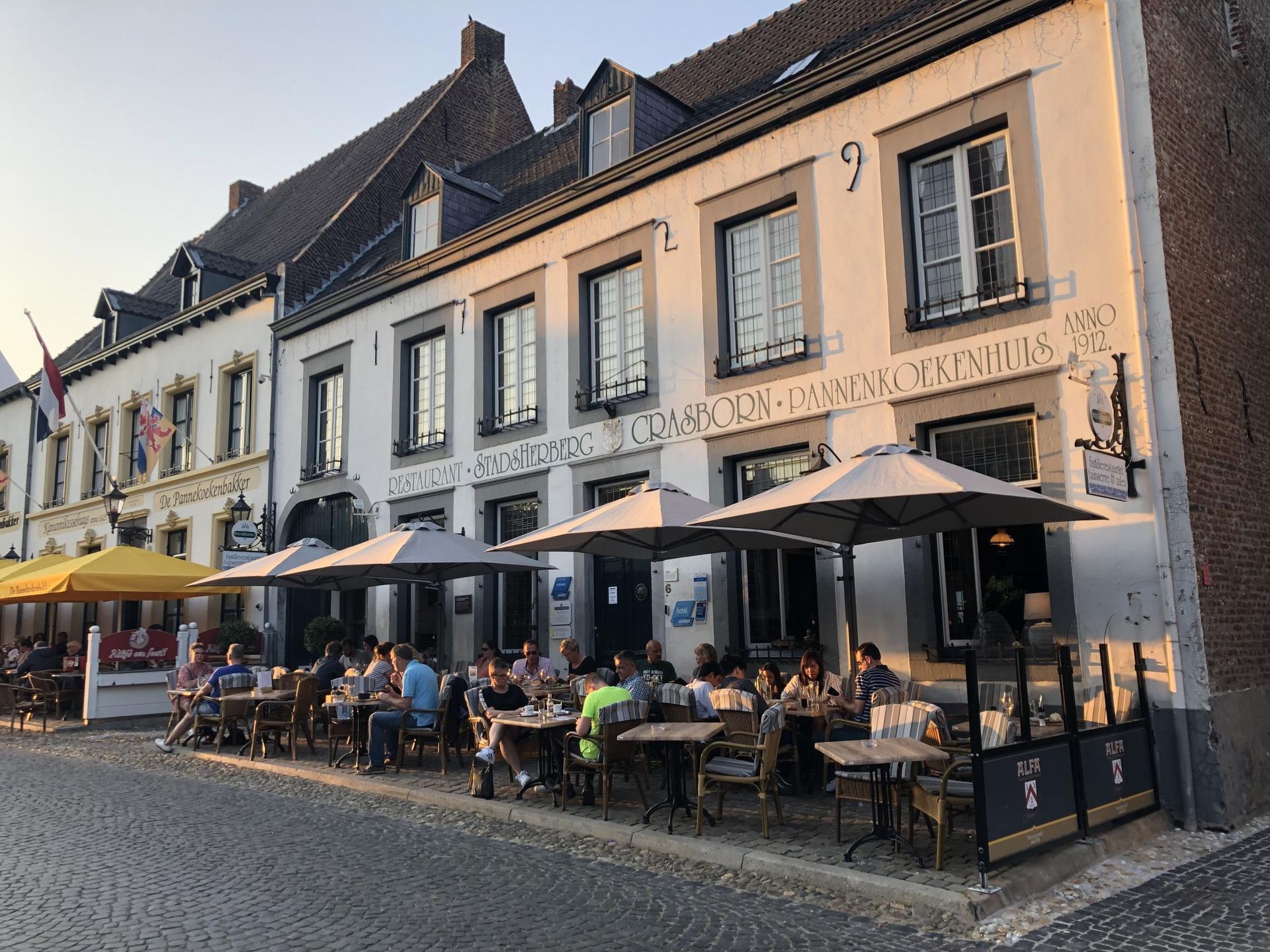 Een groep mensen zit aan tafels buiten een gebouw, met parasols.