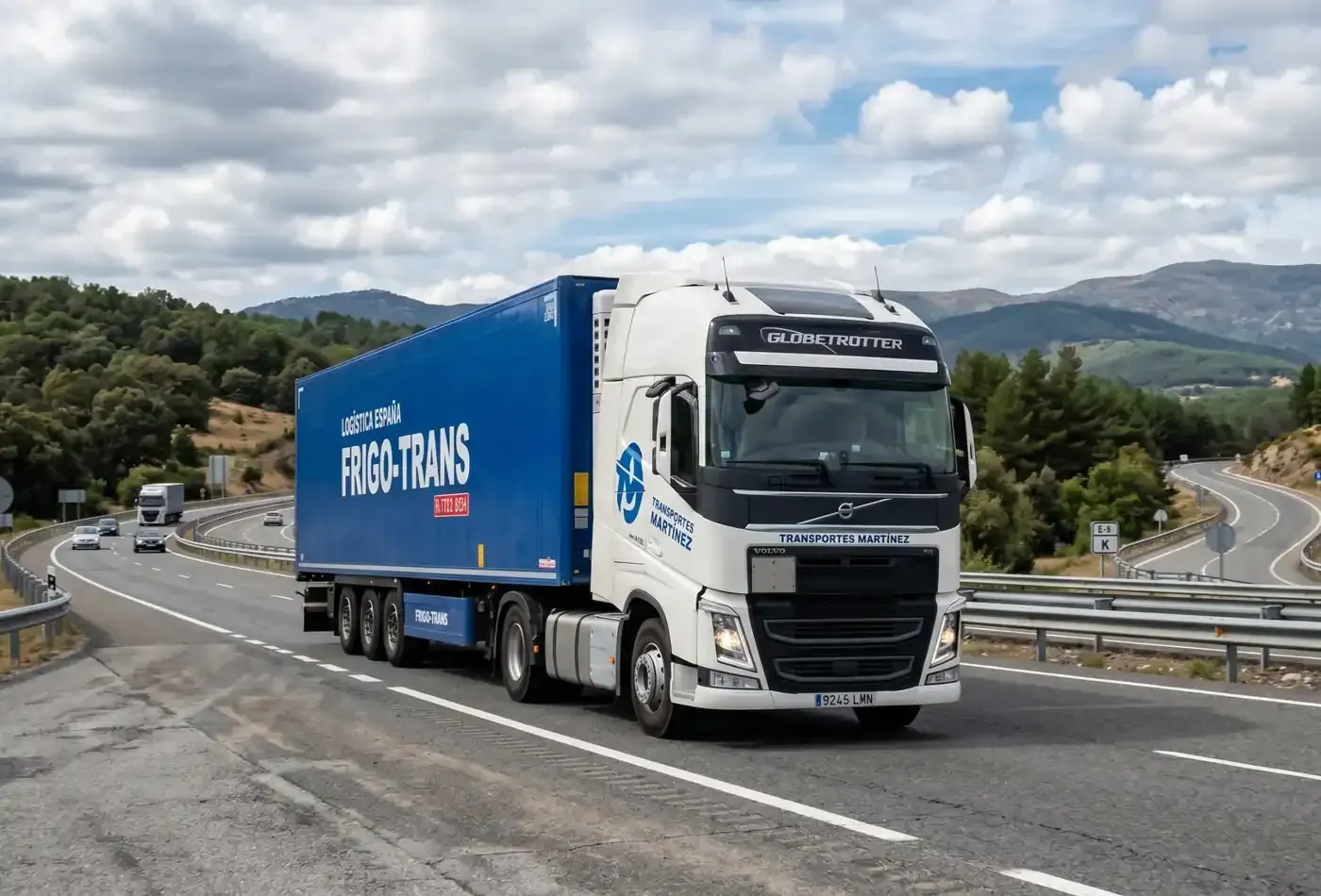 Camión de carga blanco con remolque azul circulando por una carretera de montaña sinuosa bajo un cielo nublado.