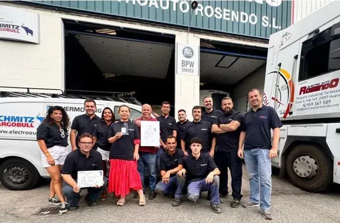 Un grupo de personas posa junto a camiones frente a un almacén, sosteniendo certificados y sonriendo.
