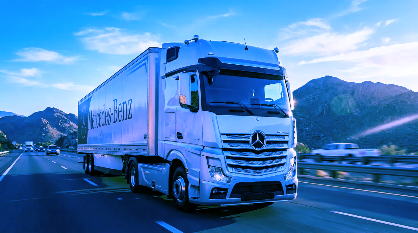 Un camión semirremolque Mercedes blanco circula por una autopista con montañas al fondo al amanecer.