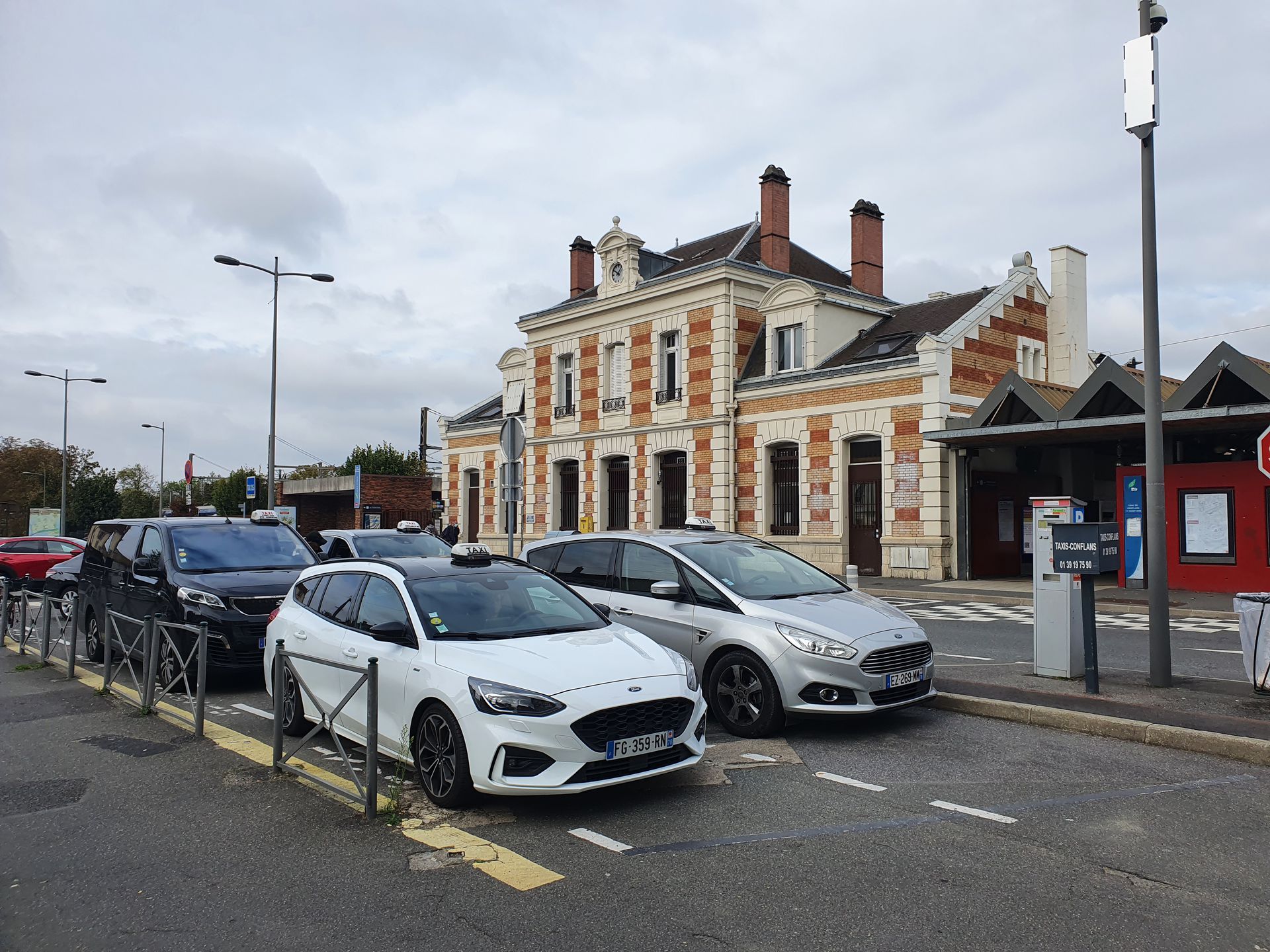 Voitures garées devant une gare en briques sous un ciel nuageux.