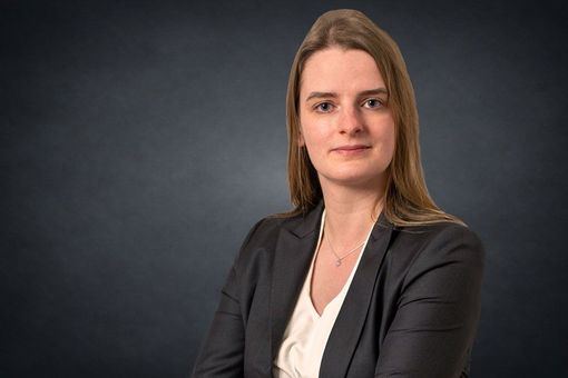 Mann in hellblauem Blazer und weißem Hemd, lächelnd vor dunkelgrauem Hintergrund.