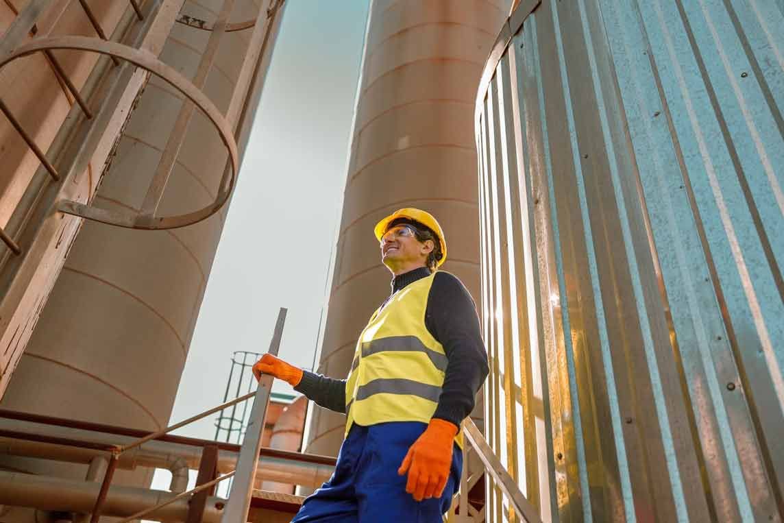Arbeiter in Sicherheitsausrüstung blickt in der Nähe von Industrietanks nach oben.