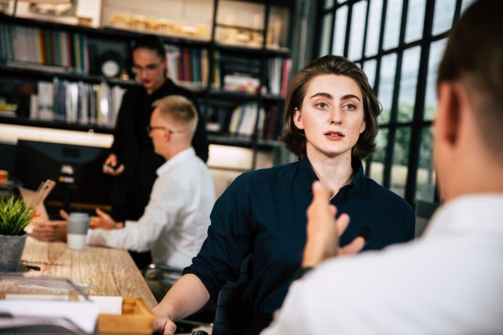 A woman gestures while speaking to a man, possibly in an office. Other people work nearby.