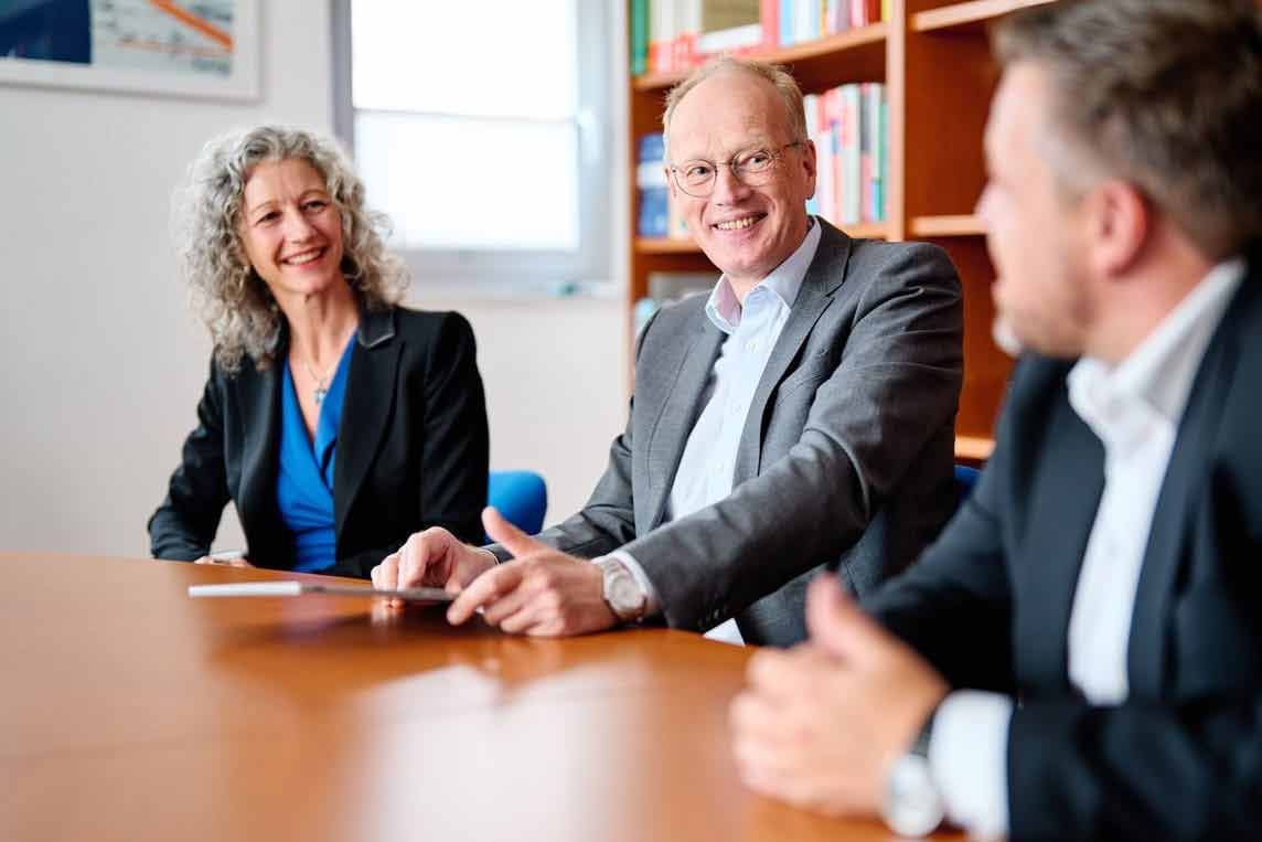 Drei Geschäftsleute sitzen lächelnd an einem Tisch. Eine Frau mit grauem Lockenhaar und blauem Oberteil, ein Mann mit Brille und grauem Anzug, ein anderer Mann mit dunklem Anzug.