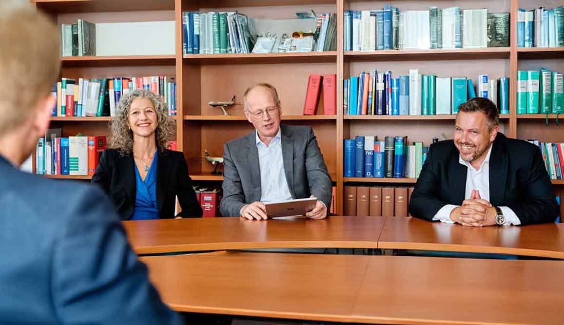 Vier Personen an einem Konferenztisch in einer Bibliothek. Drei sind sichtbar und lächeln, mit Blick auf eine Person im Vordergrund.