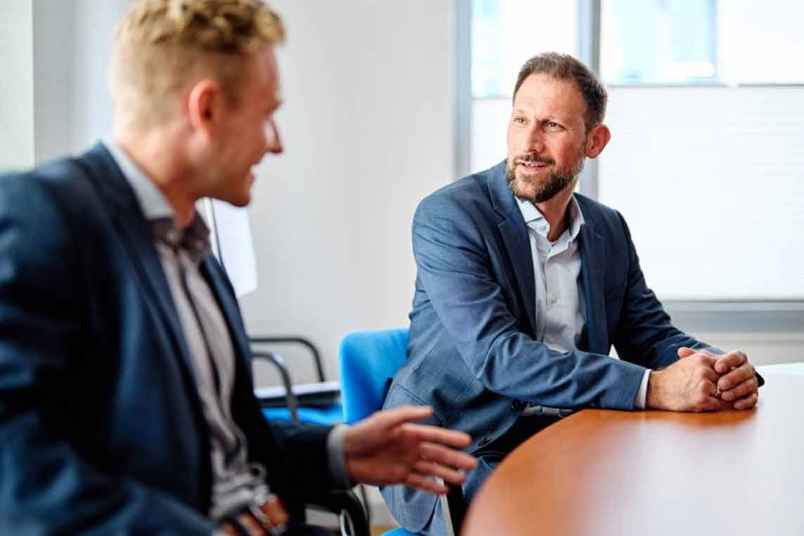 Zwei Männer in Anzügen an einem Tisch, einer spricht mit den Händen, der andere hört in einer Büroumgebung aufmerksam zu.