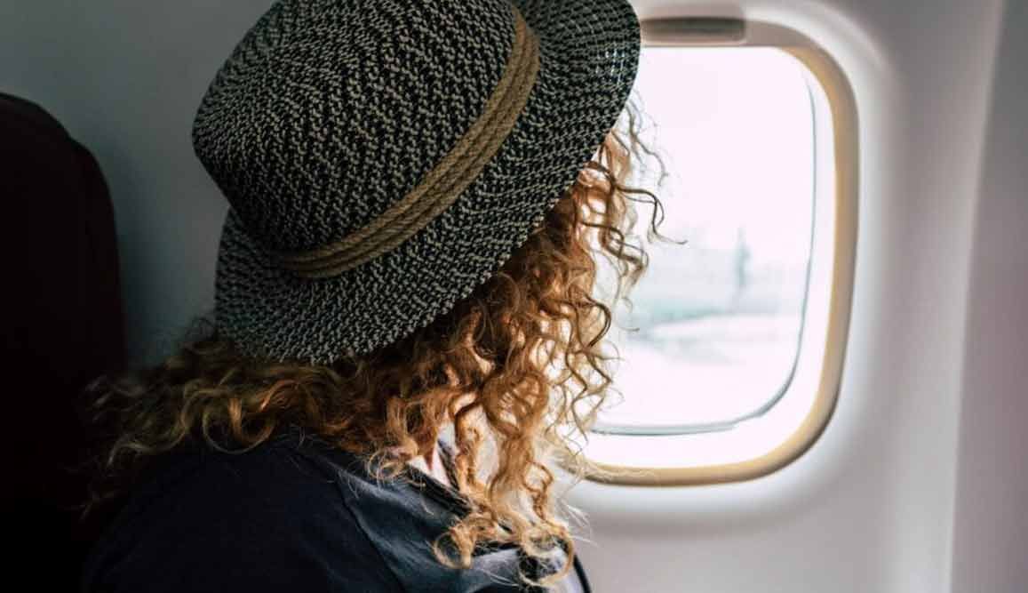 Frau mit lockigem Haar und Hut schaut aus einem Flugzeugfenster.