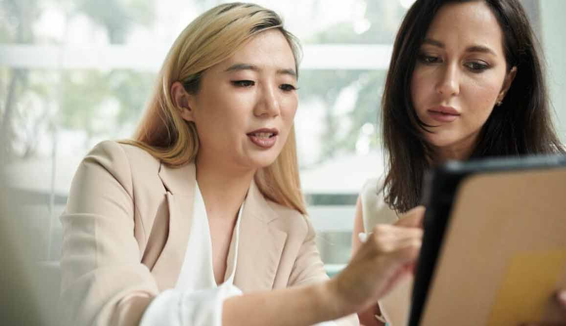 Zwei Geschäftsfrauen, eine Asiatin, eine Weiße, schauen auf ein Tablet. Die Asiatin zeigt auf den Bildschirm. Sie befinden sich in einem Büro.
