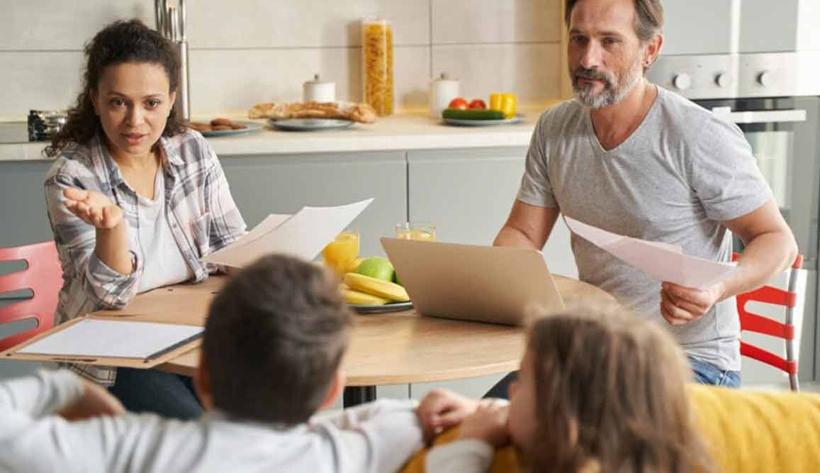 Familie sitzt am Küchentisch und bespricht Papiere. Eine Frau gestikuliert mit der Hand, ein Mann hält Papiere und zwei Kinder schauen zu.