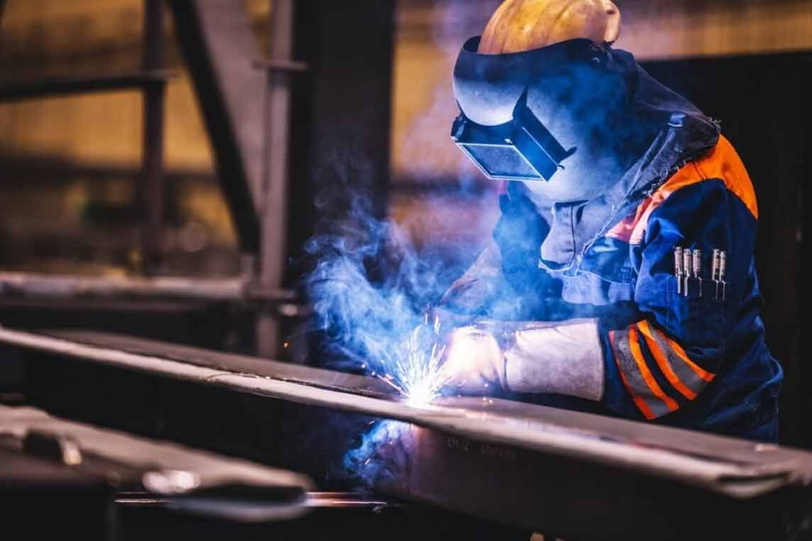 Schweißer in blauer und orangefarbener Arbeitskleidung schweißt in einer Werkstatt einen Stahlträger mit leuchtend blauen Funken.