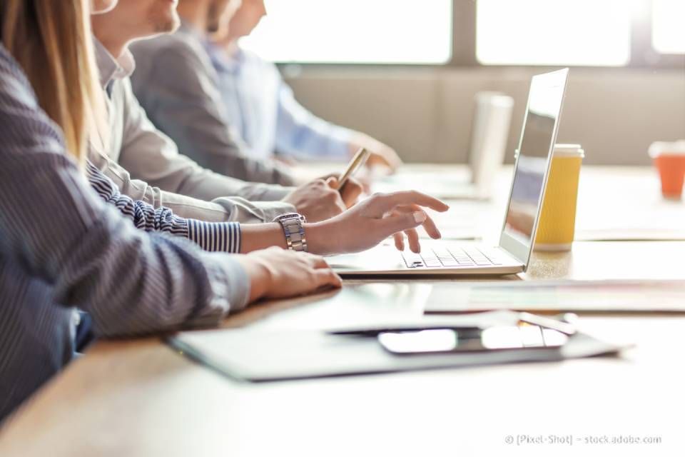 Menschen arbeiten an einem Tisch und benutzen Laptops. Die Hände einer Person tippen, die anderen sind im Hintergrund verschwommen zu sehen.