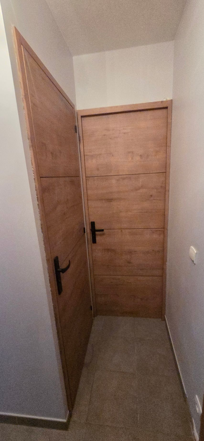 Dans une pièce gris foncé meublée d'un meuble TV, d'une plante et d'étagères, une porte pliante blanche donne sur une salle de bains lumineuse.