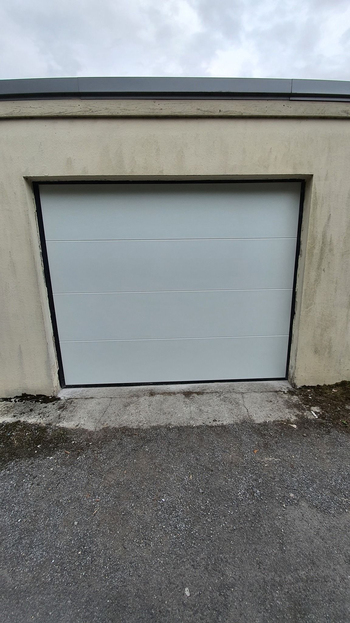 L'extérieur d'une maison moderne avec deux portes de garage grises encastrées dans un mur de pierre texturée de couleur claire, sous un ciel bleu azur.