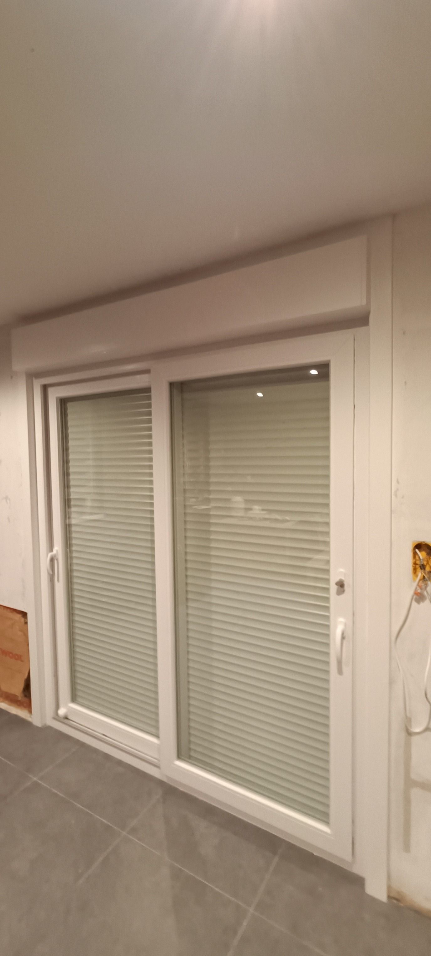 Une fenêtre gris foncé avec un volet roulant perforé partiellement abaissé, adossée à un mur sombre donnant sur un champ.