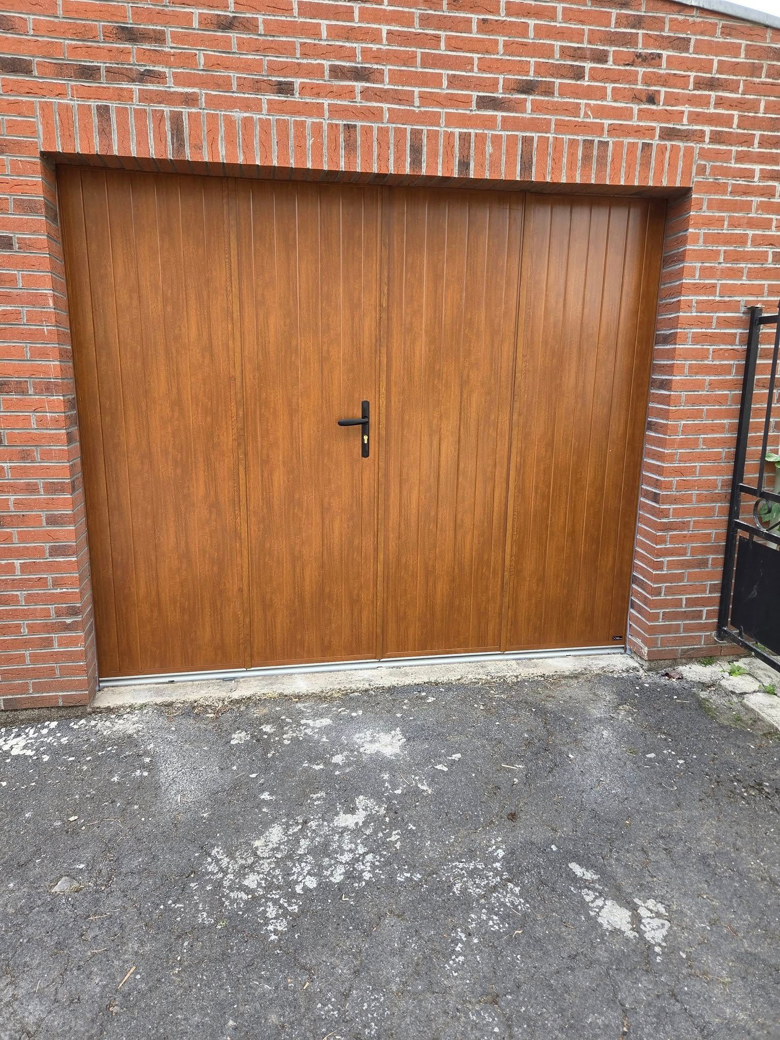Une porte de garage moderne gris foncé jouxte une porte d'entrée assortie, dotée de panneaux de verre, et deux grandes jardinières ornent une terrasse pavée.
