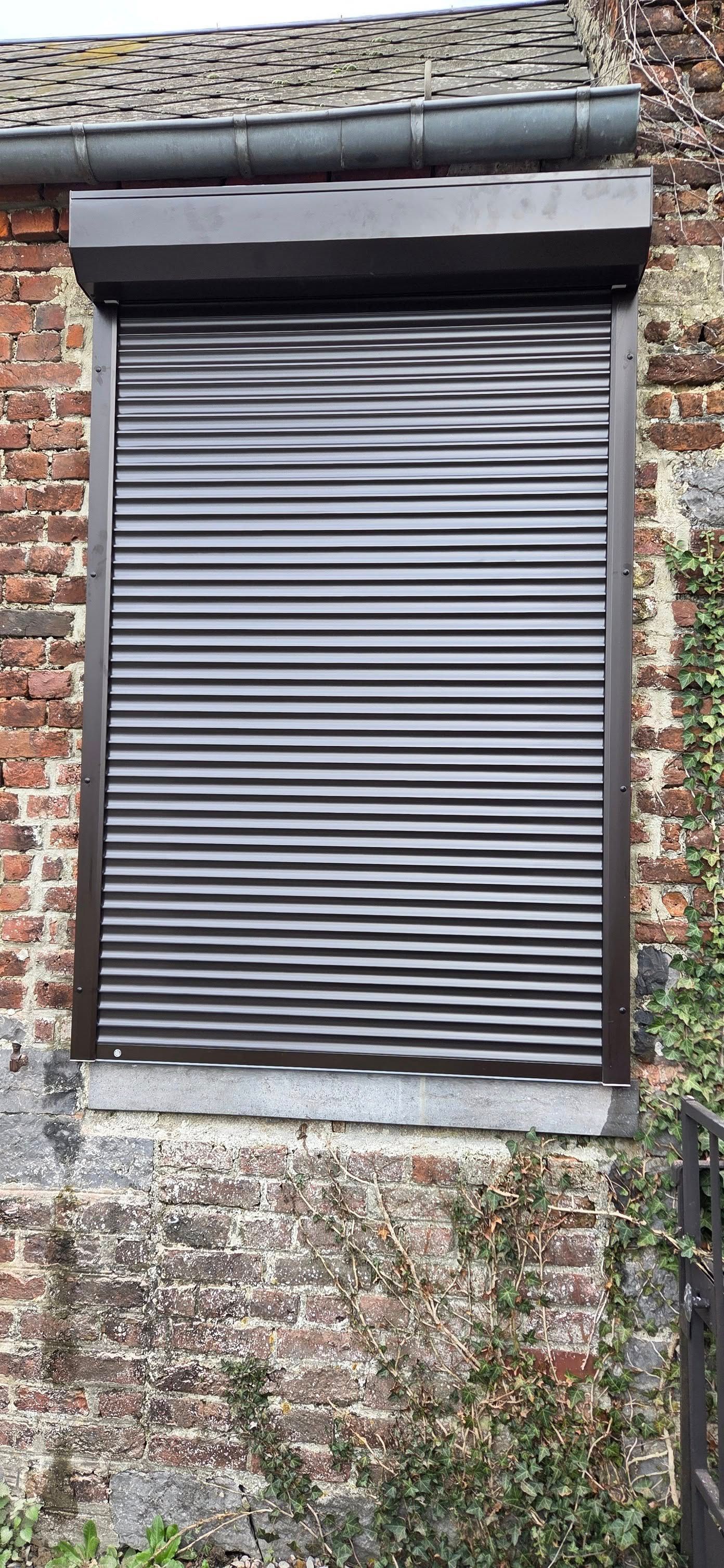 Un volet roulant de sécurité blanc et horizontal installé sur une fenêtre d'un mur extérieur gris à parois horizontales.