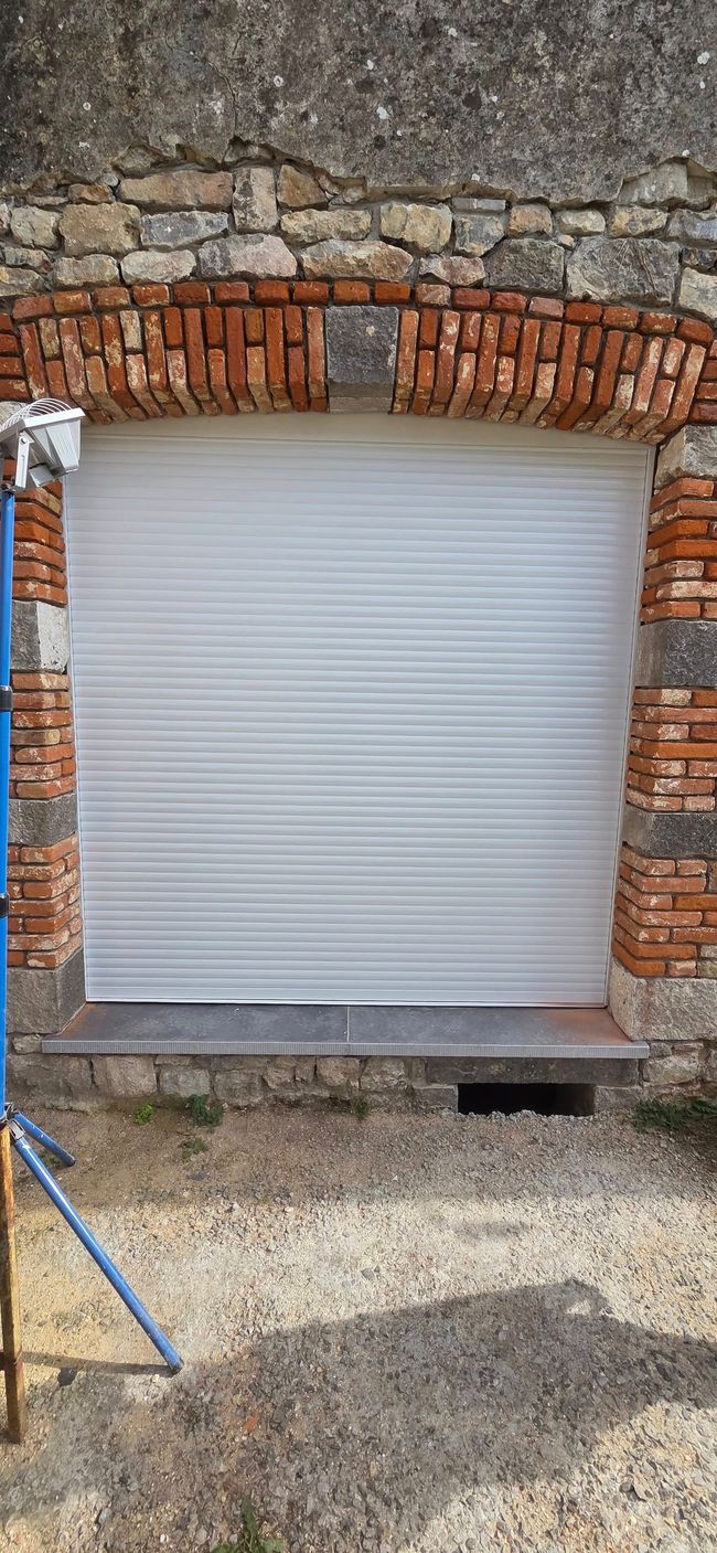 Patio d'une maison moderne doté de grands stores extérieurs rétractables sombres au-dessus de baies vitrées donnant sur un jardin verdoyant.