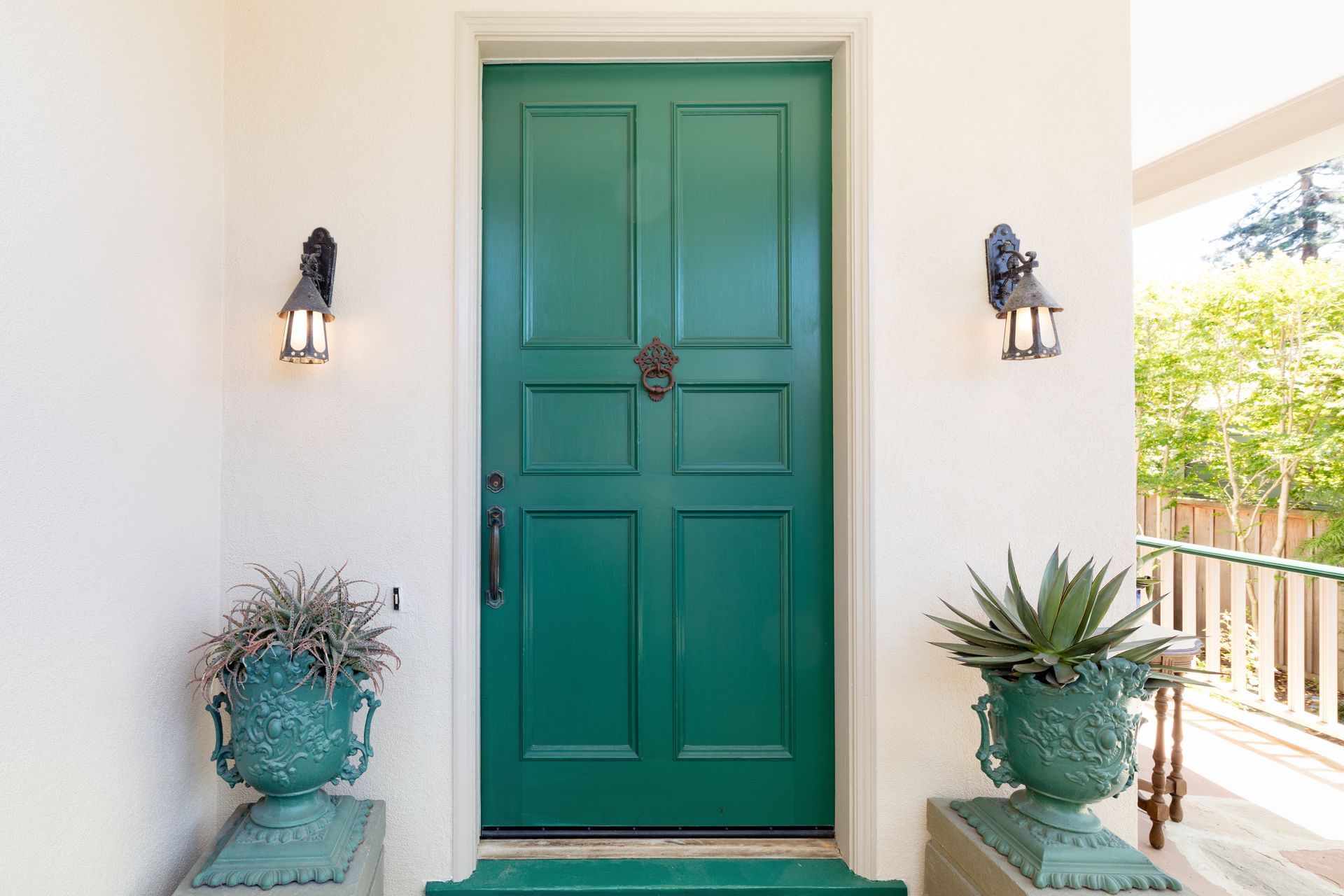Une porte d'entrée turquoise avec un heurtoir, flanquée d'appliques murales et de plantes en pot sur un porche blanc.