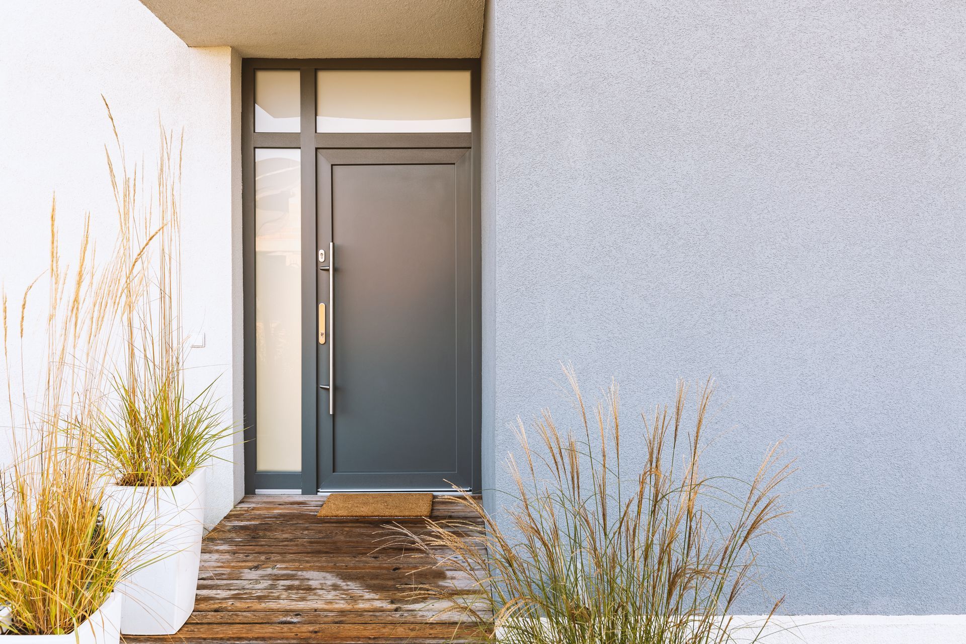 Une porte d'entrée moderne gris foncé avec une fenêtre latérale, nichée dans un mur gris texturé orné de graminées ornementales en pot.