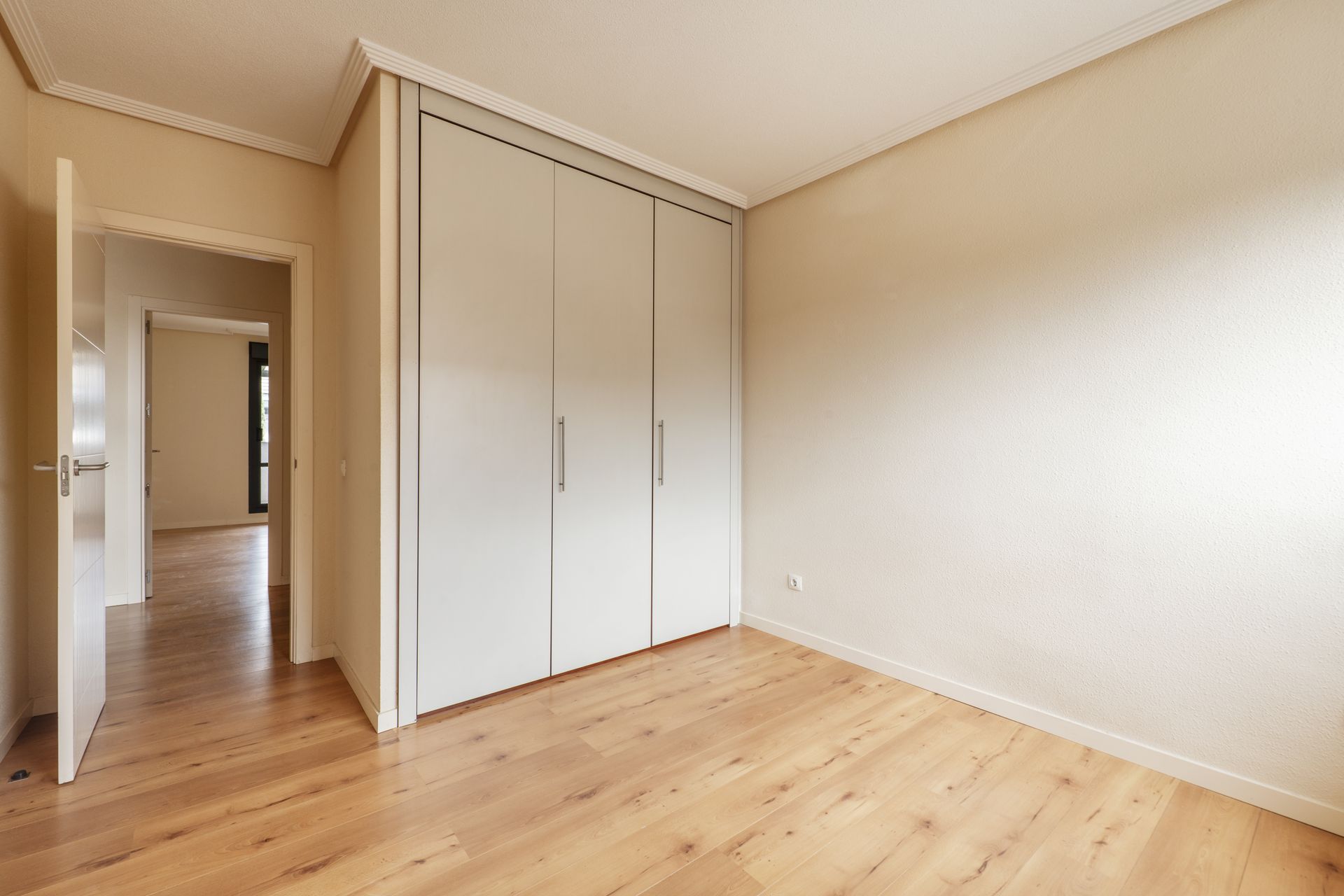 Une chambre lumineuse et vide, avec parquet clair, murs beiges et grande armoire blanche encastrée à trois portes.