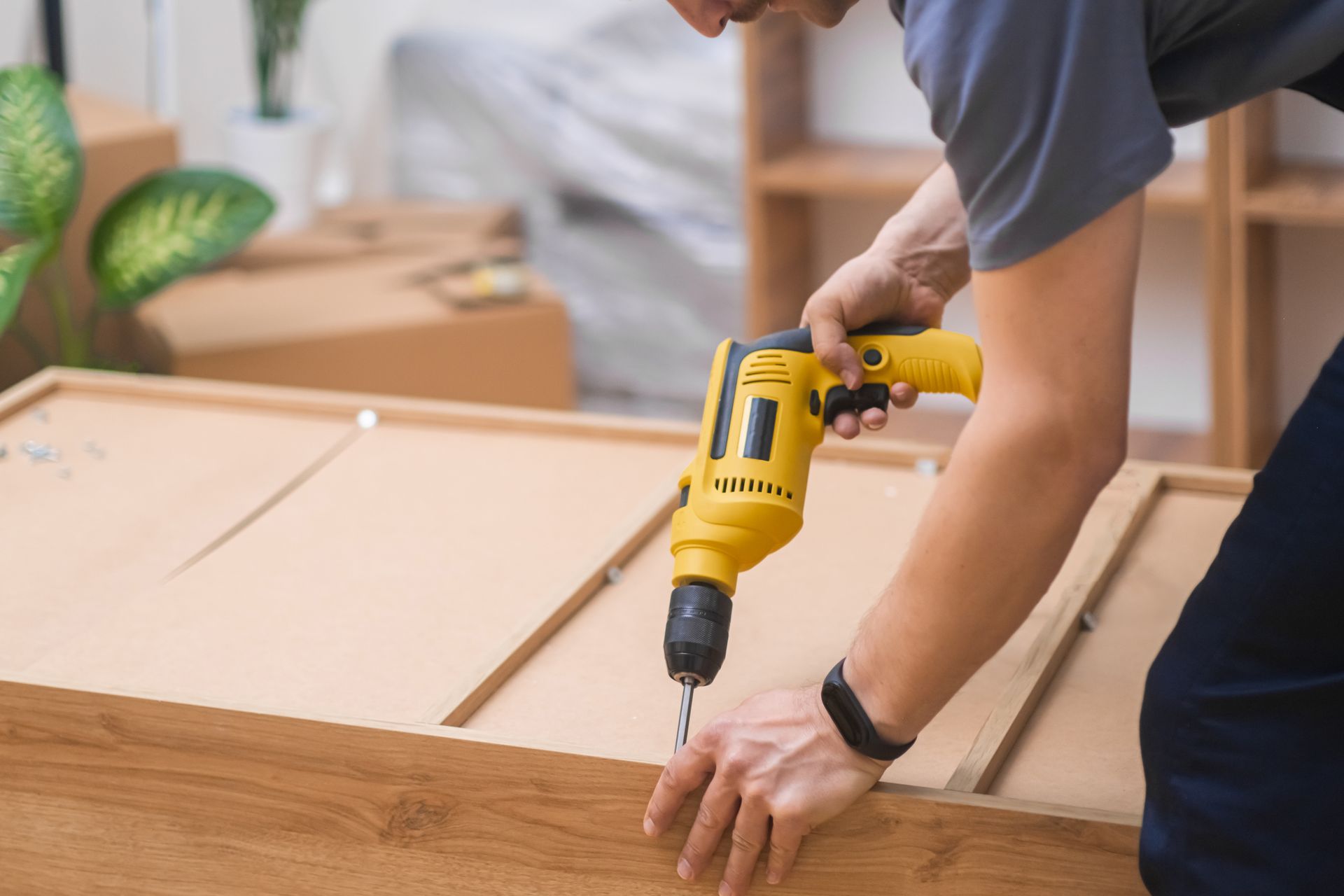 Dans un intérieur lumineux, une personne utilise une perceuse électrique jaune pour assembler un meuble en bois.